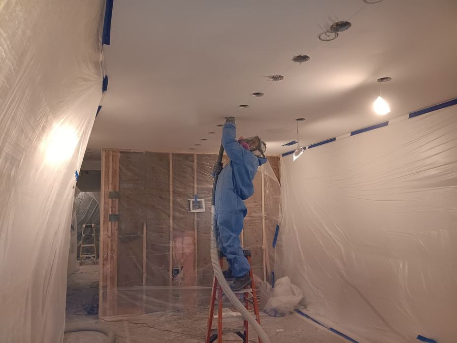 Insulation technician in protective suit spraying foam on a ceiling inside plastic containment